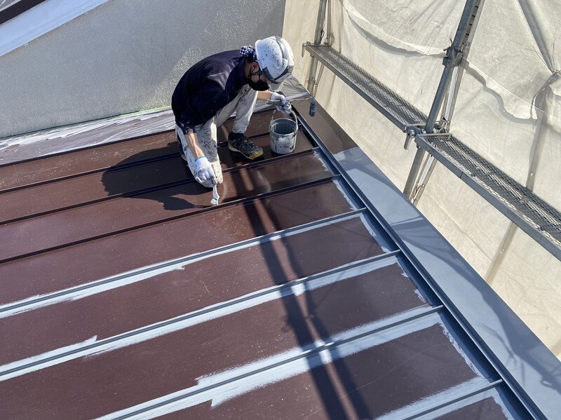 トタン屋根の狭い部分を刷毛を使って中塗り（ダメ込み）をしています。（エスケー化研 クールタイトSi CLR-106 グレー）